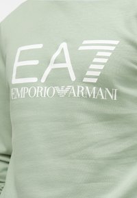 Lichtgroene sweater van katoen, met grote witte grafische tekst "EA7 EMPORIO ARMANI" en gestreepte accenten op de letters.