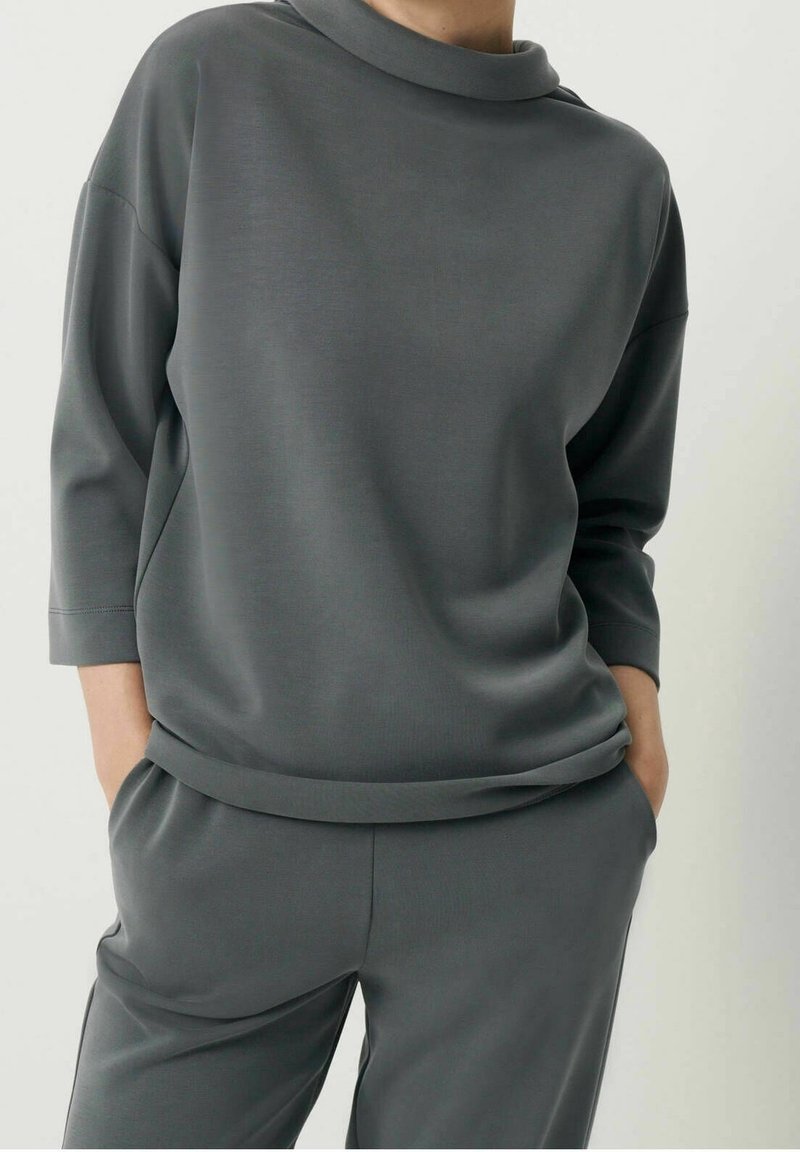 Grå sweatshirt med hög, vikt krage; tillverkad av mjukt och slätt tyg. Har trekvartslånga ärmar och en avslappnad passform. Matchas med byxor i samma färg.