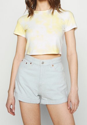 Vrouw draagt een lichtgeel en wit cropped t-shirt en lichtblauwe hooggetailleerde denim shorts met opgerolde zomen.