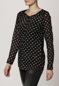 Blouse noire transparente à motif répétitif de lèvres, manches longues, boutons sur le devant et détail accent rouge à l'ourlet.