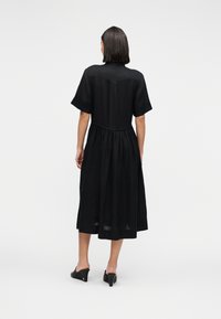 Γυναίκα με  ίσια σκούρα μαλλιά, που φοράει μαύρο μίντι φόρεμα με κοντά μανίκια και πλισέ φούστα, στέκεται με την πλάτη προς τα εμπρός σε λευκό φόντο.