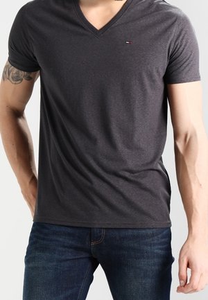 Homme portant un T-shirt gris foncé ajusté à col en V avec un petit logo sur la poitrine et un jean bleu, debout avec une main sur la hanche.