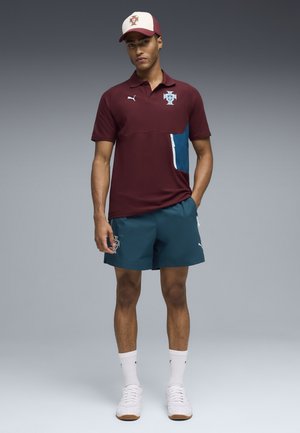 Mand stående iført en vinrød polotrøje, turkis shorts, hvide sneakers, hvide sokker og en beige og vinrød baseballkasket.