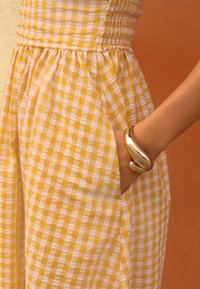 Mano nella tasca dell'abito con un braccialetto d'oro curvo, abito a quadri giallo e bianco, con uno sfondo caldo marrone e giallo.