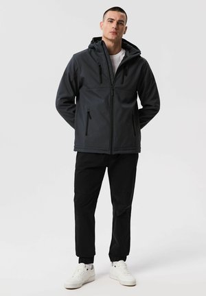 Mężczyzna w czarnej kurtce z kapturem, czarnych spodniach i białych trampkach, stoi z rękami za plecami na jednolitym jasnym tle.