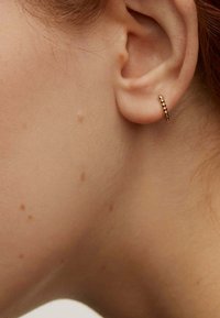 Pendiente de aro dorado con un diseño texturizado y pequeños acentos esféricos. Llevado en la oreja de un modelo con un tono de piel claro y texturizado.