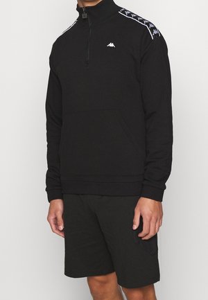 Mann trägt schwarz viertelreißverschluss Sweatshirt mit weißem Logo und schwarze Shorts vor einfarbigem Hintergrund.