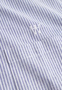 White fabric with thin blue stripes, featuring an embroidered "W" on the chest pocket. Smooth texture with a classic design.