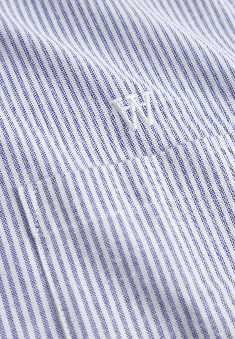 Tela blanca con finas rayas azules, que presenta una "W" bordada en el bolsillo del pecho. Textura suave con un diseño clásico.
