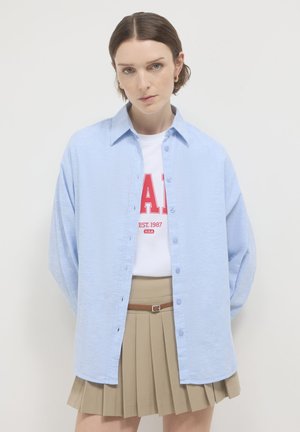 Light blue button-up shirt made of textured fabric, featuring a collar and dropped shoulders, paired with a white graphic T-shirt and beige pleated skirt.