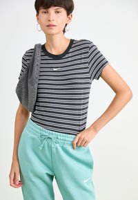 Ung kvinne som har på seg en svart og hvit stripete Nike T-skjorte og lyseblå Nike joggebukser, og bærer en grå bag over skulderen.