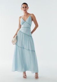 Světle modré maxi šaty s plisovanou texturou, tenkými ramínky a vrstveným designem zvýrazněným ozdobným lemem. Nošeny s bílými sandály.