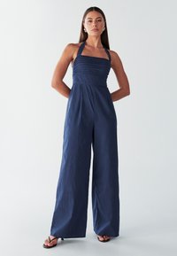 Mornarsko modra kombineza z vrvico za okoli vratu, oprijetim zgornjim delom, nabranim teksturo in širokimi hlačnicami. Izdelana iz lahkega materiala, s poudarkom na minimalističnem videzu.