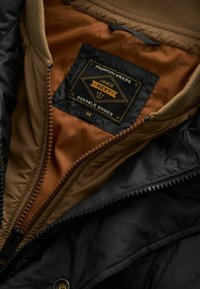 Sort og brun pufferjakke indvendigt med en brun foring, synlig lynlås og et sort mærke, der viser "Premium Grade 1982 Next Durable Goods."