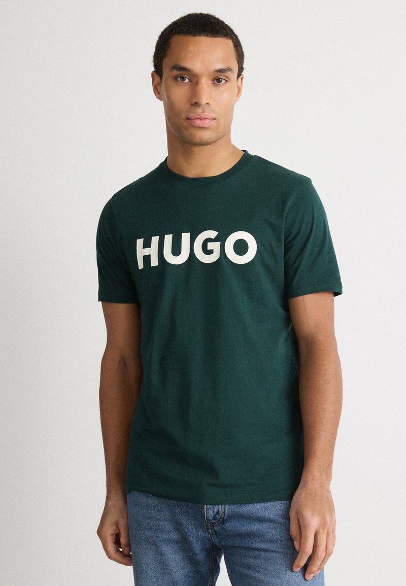 T-shirt de algodão verde escuro com mangas curtas e decote redondo. Apresenta o texto em branco em destaque "HUGO" na parte frontal.