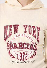 Sudadera beige con un gráfico frontal que dice "NUEVA YORK" y "GARCIA" en texto burdeos desgastado, acentuado por hojas de laurel y el año "1972."