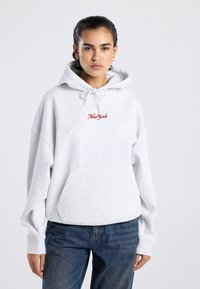 Joven con cabello oscuro lleva una sudadera gris claro con el texto "New York" en rojo y jeans azules, de pie frente a un fondo liso.