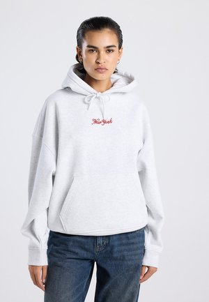 Jong persoon met donker haar draagt een lichtgrijze hoodie met de tekst "New York" in het rood en blauwe jeans, staand tegen een effen achtergrond.