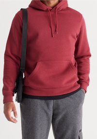 Röd hoodie gjord av mjukt material, med en ficka fram, dragsko och ribbade ärmslut; ihop med grå sweatpants och en svart väska.