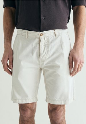 Hombre que lleva pantalones cortos beige hasta la rodilla y una camisa oscura con botones, de pie con los brazos relajados a los lados.