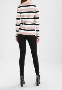 Donna che indossa una maglia a maniche lunghe a righe rosa, bianca e nera, jeans skinny neri e stivaletti neri con borchie, in piedi con le spalle rivolte.