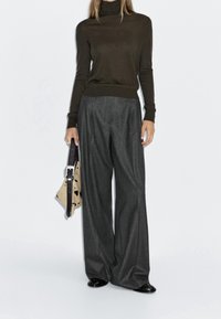 Femme portant un pull à col roulé marron foncé, un pantalon large gris foncé, des chaussures noires, tenant un sac à main beige à taches noires avec une boucle.