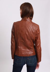 Giubbotto in pelle marrone con un design aderente, cuciture verticali e colletto alto, indossato sopra jeans in denim scuro. Texture liscia, dettagli minimi.