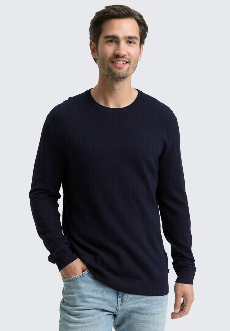 Mann mit kurzen dunklen Haaren und leichtem Stoppelschnitt trägt einen dunkelblauen langärmeligen Pullover und hellblaue Jeans, steht mit einer Hand in der Tasche.
