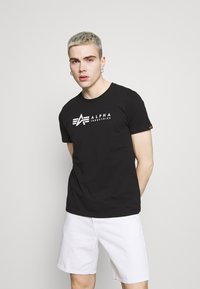 Juodas medvilnės marškinėliai su balta "Alpha Industries" logotipu per krūtinę. Papildyti šviesių spalvų šortais. Paprastas dizainas.