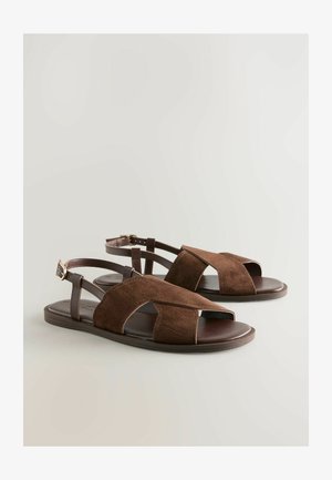 Brūni zamšādas sandales ar atvērtu purngali, krusta siksnām un regulējamu potītes sprādzi. Gluda brūna zole ar minimālu saķeri. Plakans dizains.