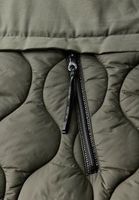 Tkanina w kolorze oliwkowym o pikowanej strukturze, z gładką fakturą, z czarnym zamkiem błyskawicznym na środku oraz stworzonymi za pomocą haftu okrągłymi wzorami.