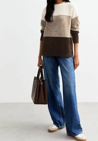 Gebreide trui met beige, taupe en bruine horizontale strepen; gecombineerd met wijde blauwe spijkerbroeken; een bruine handtas vasthoudend.