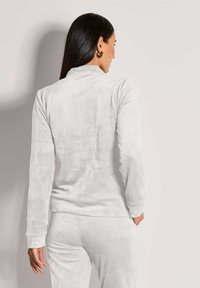 Vrouw met lang donker haar in een witte velours lange mouwen top en bijpassende broek, met haar rug naar ons gekeerd en één hand in de zak.