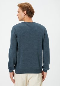 Dunkelblauer Pullover mit strukturiertem Strick, Rundhals, langen Ärmeln; gerippte Bündchen und Saum, getragen über hellen Hosen.