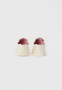 Des baskets blanches avec une texture lisse et un accent en velours rose au talon. Forme arrondie, design minimaliste, et semelle en caoutchouc épaisse.