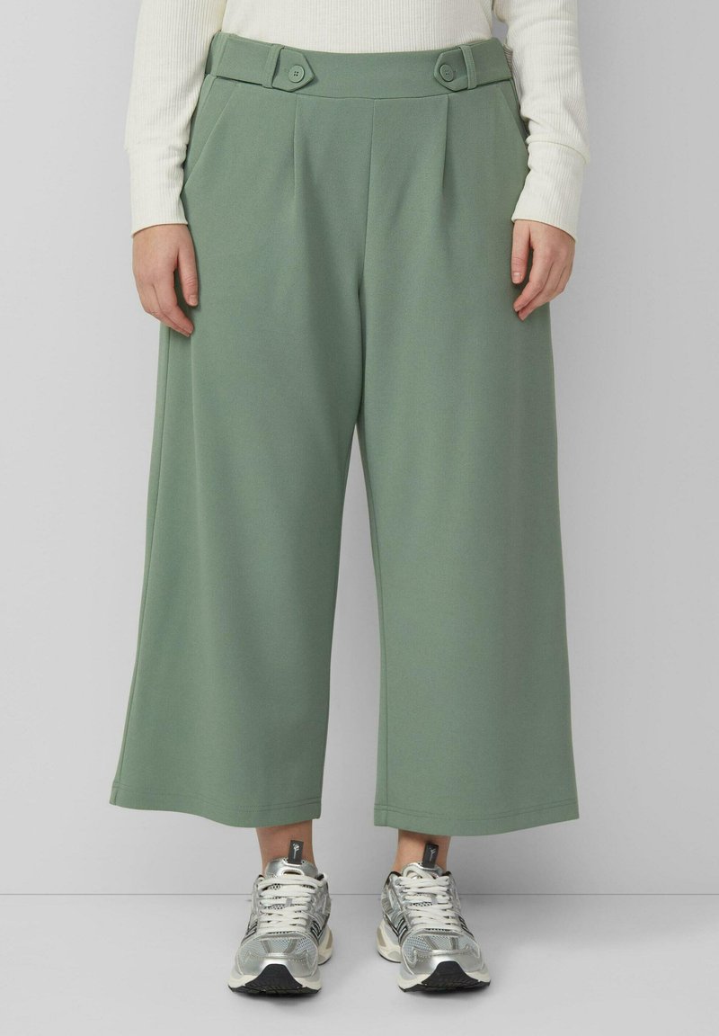 Person trägt locker sitzende, salbeigrüne, knöchellange Hose mit Knopfdetails und silbernen Sneakern, steht vor einem schlichten hellgrauen Hintergrund.