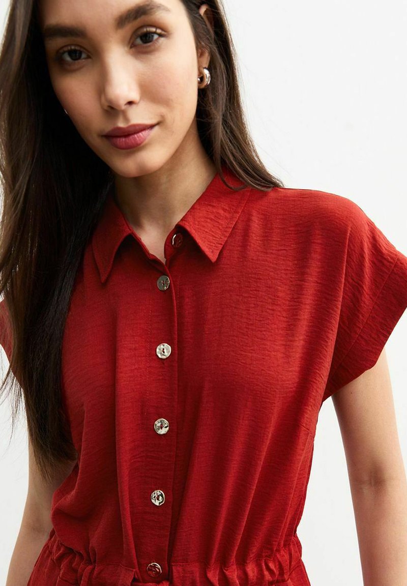 Vestido rojo de botones, hecho de una tela texturizada, con mangas cortas, un cuello con solapas y botones plateados en la parte delantera.