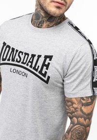 Gray cotton t-shirt featuring black "LONSDALE LONDON" graphic, round neckline, and black striped accents on the shoulders.