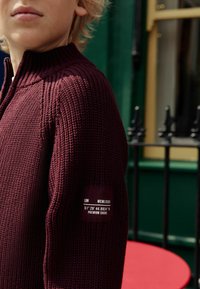 Pull en maille bourgogne avec col haut, texture côtelée et étiquette noire affichant des coordonnées et du texte sur la manche gauche.