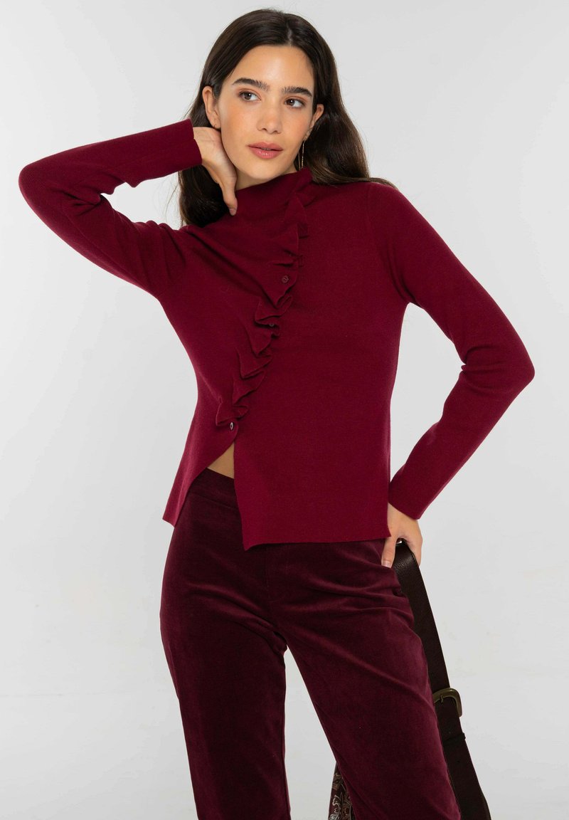 Suéter burdeos de punto con cuello alto, detalles de volantes en un lado y botones. Combinado con pantalones de terciopelo burdeos.