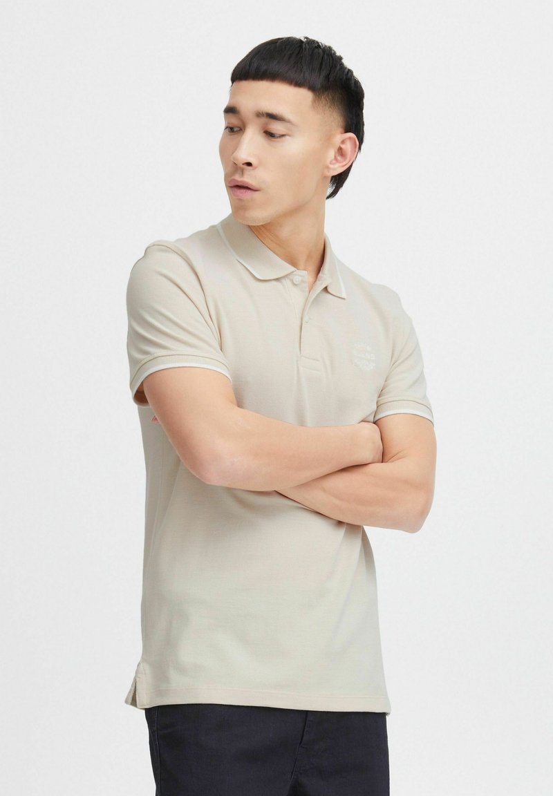Beige polo skjorte med krage, korte ermer, og et logo på brystet. Glatt stoff med kontrasterende kanting, kombinert med mørke bukser.