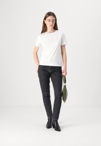 T-shirt bianco in cotone con scollo rotondo, abbinata a jeans slim-fit scuri. La modella tiene una borsa verde e indossa stivaletti neri alla caviglia.