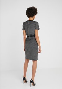 Femme aux cheveux courts et bouclés portant une robe rayée noire et blanche jusqu'aux genoux avec une ceinture noire et des talons hauts noirs, debout contre un mur blanc.