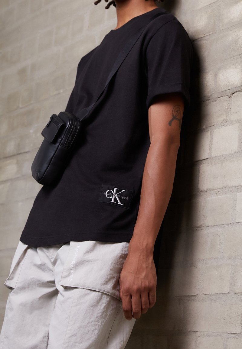 Person med svart Calvin Klein-t-shirt och ljusa cargoshorts med Calvin Klein-logotyp, bärande en svart axelremsväska, lutar sig mot en beige tegelvägg.