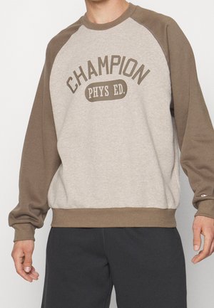 Personne portant un sweat-shirt beige et marron à manches longues avec "CHAMPION PHYS ED." imprimé sur le devant et un pantalon foncé.