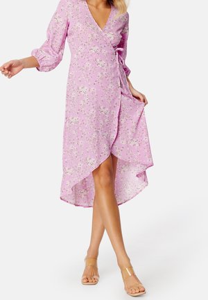 Femme portant une robe portefeuille rose à fleurs avec des manches longues et un ourlet asymétrique, associée à des talons ouverts à lanières transparentes.