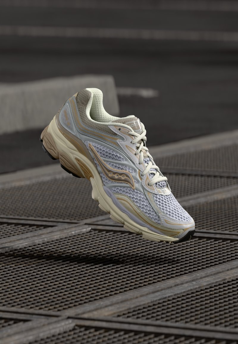 Λευκά και ανοιχτόχρωμα γκρι αθλητικά παπούτσια με δικτυωτό επάνω μέρος, καφέ τόνους και μαξιλαρωτή σόλα. Διαθέτει περίπλοκα μοτίβα αερισμού και κλείσιμο με κορδόνια.