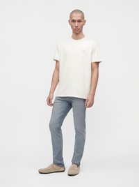 Άνδρας στέκεται φορώντας μια απλή off-white κοντομάνικη μπλούζα, ανοιχτό μπλε τζιν και μπεζ παπούτσια χωρίς κορδόνια σε λευκό φόντο.