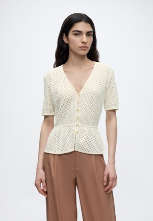 Jonge vrouw met lang donker haar draagt een crèmekleurige, korte mouwen, geknoopte gehaakte top en losse bruine broek tegen een effen achtergrond.