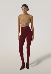 Kvinna med slickat hår iförd en beige bandeau topp, ribbade vinröda högmidjade leggings och svarta högklackade skor som står mot en ljus bakgrund.